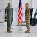 Two U.S. Air Force personnel are participating in what appears to be a formal military ceremony, likely a swearing-in or reenlistment, inside a hangar facility. The officer on the left, wearing an olive green flight suit, has his right hand raised in an oath gesture, while the airman on the right in OCP (Operational Camouflage Pattern) uniform stands at attention. An American flag on a ceremonial stand is positioned between them.