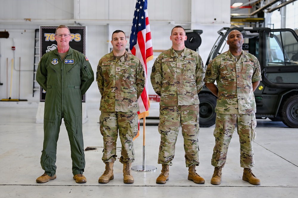 Four U.S. Air Force personnel are posed for a formal group photo inside what appears to be an aircraft maintenance hangar. One individual on the left wears an olive green flight suit, while the other three wear OCP (Operational Camouflage Pattern) uniforms. An American flag on a stand is positioned between the second and third individuals.