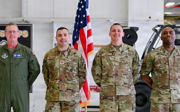 Four U.S. Air Force personnel are posed for a formal group photo inside what appears to be an aircraft maintenance hangar. One individual on the left wears an olive green flight suit, while the other three wear OCP (Operational Camouflage Pattern) uniforms. An American flag on a stand is positioned between the second and third individuals.
