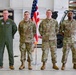 Four U.S. Air Force personnel are posed for a formal group photo inside what appears to be an aircraft maintenance hangar. One individual on the left wears an olive green flight suit, while the other three wear OCP (Operational Camouflage Pattern) uniforms. An American flag on a stand is positioned between the second and third individuals.
