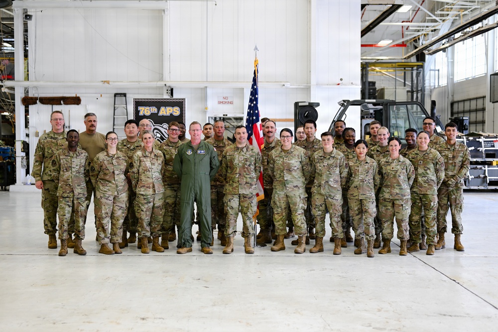 A group of approximately 25-30 military personnel pose for a formal unit photo inside what appears to be a military hangar or maintenance facility. The majority are wearing OCP (Operational Camouflage Pattern) uniforms, with one individual in a green flight suit standing centrally. A banner reading '76th APS' and an American flag are visible in the background.