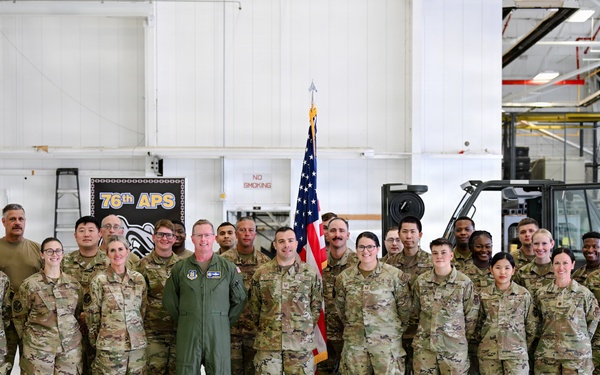A group of approximately 25-30 military personnel pose for a formal unit photo inside what appears to be a military hangar or maintenance facility. The majority are wearing OCP (Operational Camouflage Pattern) uniforms, with one individual in a green flight suit standing centrally. A banner reading '76th APS' and an American flag are visible in the background.