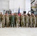 A group of approximately 25-30 military personnel pose for a formal unit photo inside what appears to be a military hangar or maintenance facility. The majority are wearing OCP (Operational Camouflage Pattern) uniforms, with one individual in a green flight suit standing centrally. A banner reading '76th APS' and an American flag are visible in the background.