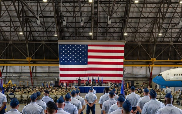 A formal military ceremony is taking place inside a large aircraft hangar, with hundreds of United States Air Force personnel assembled in formation facing a stage. A large American flag serves as the backdrop for the ceremony, which appears to be a change of command, promotion, or official military event. Two military aircraft, including what appears to be a United States government or Air Force transport aircraft, are visible inside the hangar.