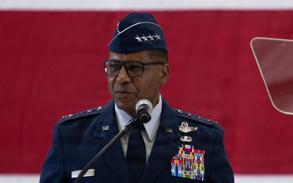 A senior U.S. Air Force officer in dress blue uniform stands at a wooden podium delivering a speech. The officer wears a service cap with four stars, indicating the rank of General, and displays an extensive array of medals and ribbons on their uniform. An American flag is visible in the background, suggesting a formal military ceremony.