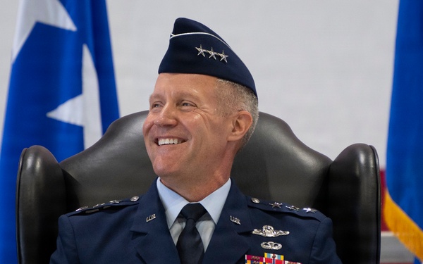 A United States Air Force officer wearing a service dress uniform is seated in a black leather chair, smiling broadly. The name tag reads 'TULLEY' and the officer wears three stars on their service cap, indicating the rank of Lieutenant General. The setting appears to be a formal military ceremony with blue and white star flags visible in the background.