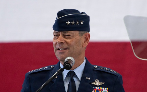 A senior U.S. Air Force officer wearing a four-star general rank insignia is speaking at a podium during what appears to be an official military ceremony. He is dressed in the Air Force Service Dress uniform with numerous ribbons, badges, and a garrison cap. A large American flag is displayed in the background.