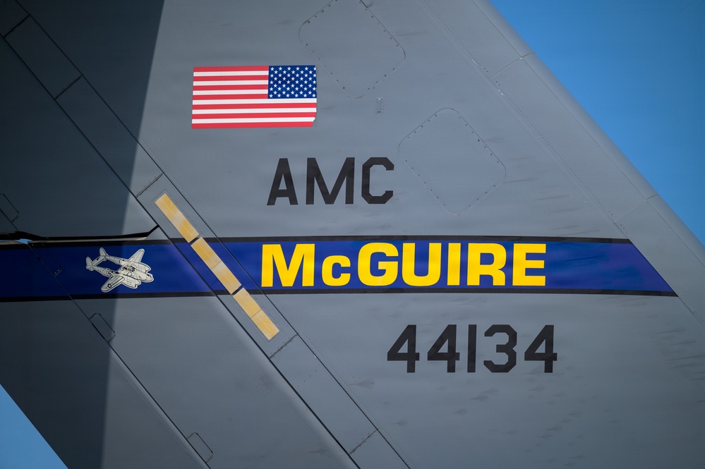 Close-up photograph of the tail section of a U.S. Air Force military aircraft assigned to McGuire Air Force Base. The tail displays markings indicating it belongs to Air Mobility Command (AMC), with the tail number 44134 and a blue and yellow 'McGuire' banner. An American flag decal and a small aircraft silhouette emblem are also visible on the tail fin.
