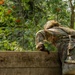 A soldier in full camouflage uniform and tactical gear is scaling or climbing over a wooden wall obstacle, likely part of a military training course. The individual is wearing a camouflage helmet, backpack, and gloves while navigating the wooden barrier. The setting is a lush, green forested environment with visible red flowering trees in the background.