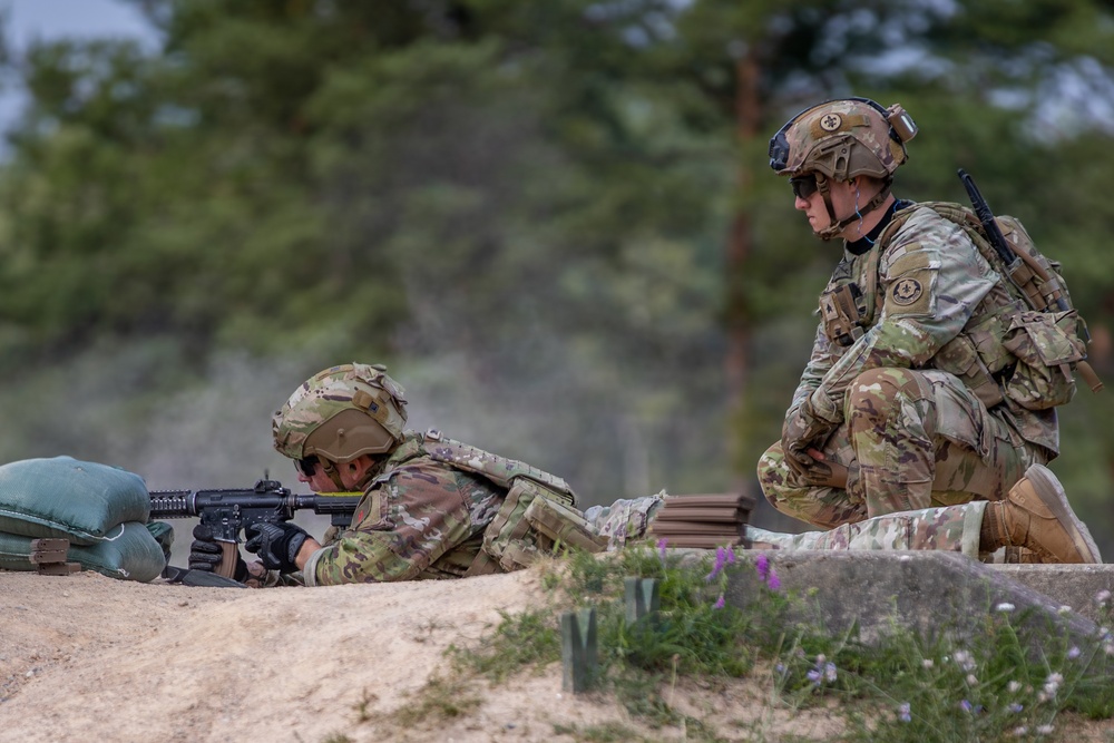 Two soldiers in MultiCam/OCP camouflage uniforms are positioned at what appears to be a firing range or defensive position. One soldier is in a prone firing position aiming a rifle, while the second soldier is crouching nearby in an observing or supervising role. The setting appears to be an outdoor training environment with sandy terrain and a treeline in the background.