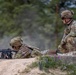 Two soldiers in MultiCam/OCP camouflage uniforms are positioned at what appears to be a firing range or defensive position. One soldier is in a prone firing position aiming a rifle, while the second soldier is crouching nearby in an observing or supervising role. The setting appears to be an outdoor training environment with sandy terrain and a treeline in the background.