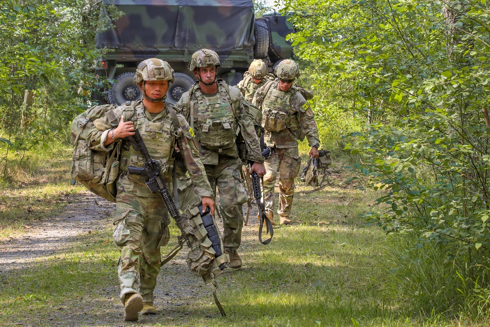 Four soldiers in U.S. Army Operational Camouflage Pattern (OCP) uniforms are moving along a dirt trail through a wooded area. They are fully equipped with tactical gear, body armor, helmets, and weapons. A military truck with a camouflage tarp cover is visible in the background.