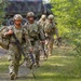 Four soldiers in U.S. Army Operational Camouflage Pattern (OCP) uniforms are moving along a dirt trail through a wooded area. They are fully equipped with tactical gear, body armor, helmets, and weapons. A military truck with a camouflage tarp cover is visible in the background.