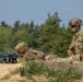 Two soldiers in multicam camouflage uniforms are positioned at what appears to be a military firing range or training position. One soldier is in a prone firing position behind sandbags aiming a rifle, while the second soldier is kneeling and observing. The setting includes a sandy berm with concrete barriers and a forested background.