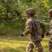 Two soldiers in military camouflage uniforms are running through a wooded outdoor area. Both wear tactical helmets with communication headsets and carry backpacks, with yellow identification patches visible on their shoulders labeled 'M5' and 'M3'. The image uses motion blur on the background, suggesting a panning camera technique to emphasize movement.