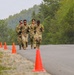 Four military personnel in camouflage uniforms are running together in formation along a paved road marked with orange traffic cones. They are wearing tactical gear including gloves and sunglasses, and appear to be participating in a timed physical fitness event or training exercise. The setting is a rural or base environment surrounded by dense green trees.