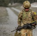 A U.S. Army soldier in full Operational Camouflage Pattern (OCP) uniform and gear is standing on or near a dirt road in a rural training environment. The soldier is looking downward while holding an M4 carbine rifle with what appears to be a training attachment. Visible name tape reads 'JOHNSON' and a yellow 'M5' marker tag is attached to the left shoulder area.