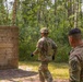Two soldiers in MultiCam/OCP camouflage uniforms are observed at what appears to be a military training course in a forested area. One soldier wearing a helmet and tactical vest is moving toward a wooden wall obstacle, while a second soldier observes from the foreground. The setting suggests a field training exercise or obstacle course evaluation.