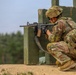 A soldier in full combat gear is positioned in a crouching stance behind a wooden barrier, aiming an assault rifle. The soldier is wearing MultiCam/OCP camouflage uniform, tactical vest, and ballistic helmet. The setting appears to be a military training range based on the constructed wooden barricade and sandy terrain.