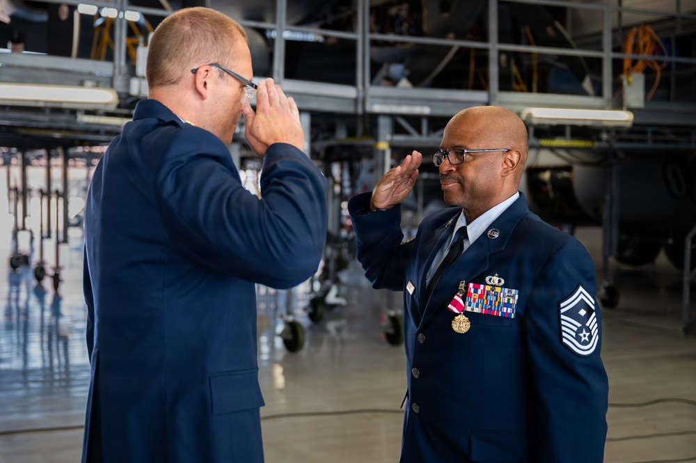 Two U.S. Air Force personnel in dress blue uniforms are engaged in what appears to be a formal military ceremony inside an aircraft hangar. The senior enlisted member on the right, identifiable by his Chief Master Sergeant rank insignia and multiple service ribbons and medals, is rendering a salute. The individual on the left appears to be photographing or filming the ceremony.
