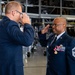 Two U.S. Air Force personnel in dress blue uniforms are engaged in what appears to be a formal military ceremony inside an aircraft hangar. The senior enlisted member on the right, identifiable by his Chief Master Sergeant rank insignia and multiple service ribbons and medals, is rendering a salute. The individual on the left appears to be photographing or filming the ceremony.