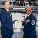 Two United States Air Force personnel are posing together in an aircraft hangar while jointly holding an official certificate or commendation document. The individual on the right, a Senior Non-Commissioned Officer with extensive service ribbons and medals, appears to be receiving or presenting the award. The setting is a large maintenance hangar with aircraft equipment and scaffolding visible in the background.