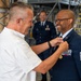 A military ceremony taking place inside an aircraft hangar where a civilian in a white shirt is pinning a medal or insignia onto a U.S. Air Force senior enlisted member. The uniformed individual appears to be a Chief Master Sergeant based on visible rank insignia, and both individuals are smiling warmly. Several other Air Force personnel in dress blues are visible in the background observing the ceremony.