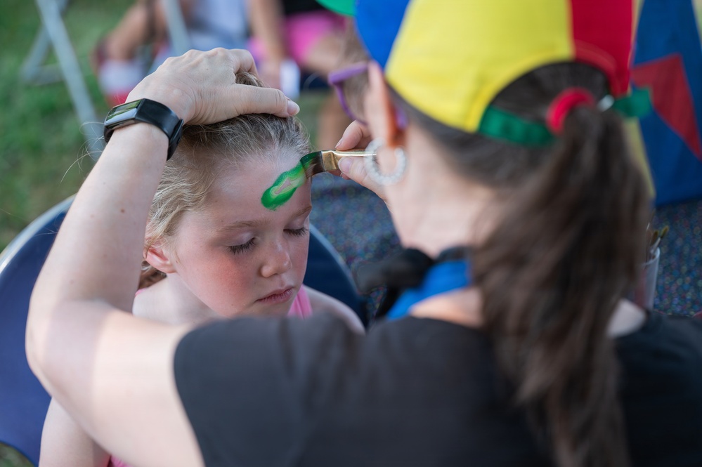 A face painter wearing a colorful jester-style hat is applying green paint to a young blonde girl's forehead using a brush. The artist steadies the child's head with one hand while carefully painting a design. The scene appears to take place at an outdoor festival or community event.