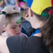 A face painter wearing a colorful jester-style hat is applying green paint to a young blonde girl's forehead using a brush. The artist steadies the child's head with one hand while carefully painting a design. The scene appears to take place at an outdoor festival or community event.