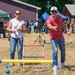 Two adults are playing ladder toss (also known as ladder ball) at an outdoor community or military event on a sunny day. A man in a red Nike polo shirt and white cap is mid-throw, releasing a bolo (two balls connected by a string), while a woman in a white top and jeans watches and holds her own bolo. A large crowd is gathered in the background near tents and structures.