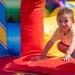 A young child, approximately 2-4 years old, is playing inside a colorful inflatable bounce house. The child is crouching and leaning forward with hands pressed on the red inflatable surface, looking directly at the camera with a slight smile. The image captures a typical recreational play setting, likely at an outdoor event or party.