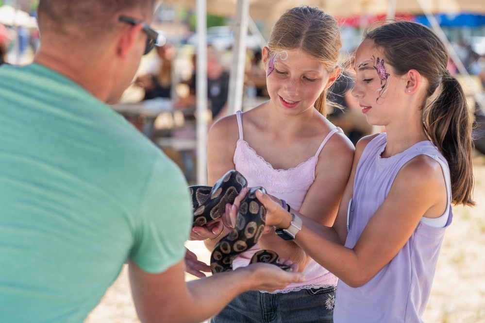 Two young girls with decorative face paint are holding a ball python snake at what appears to be an outdoor fair or festival. An adult male in a green shirt is assisting them, likely a handler presenting the snake for interaction. The setting includes tents and crowds visible in the blurred background.