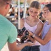Two young girls with decorative face paint are holding a ball python snake at what appears to be an outdoor fair or festival. An adult male in a green shirt is assisting them, likely a handler presenting the snake for interaction. The setting includes tents and crowds visible in the blurred background.