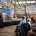 A formal ceremony is taking place inside a large military hangar, with a civilian speaker addressing an audience from a wooden podium on a raised stage. Behind the speaker, a military officer in camouflage uniform is seated, and a blue neon backdrop displays what appears to be the outline of West Virginia with a military aircraft. Multiple military service flags are visible on the right side of the stage area.