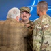 A military decoration ceremony is taking place in front of a blue backdrop featuring a lightning bolt and aircraft silhouette design. A civilian in a plaid jacket is pinning a medal onto a uniformed service member wearing OCP (Operational Camouflage Pattern) uniform and cap, while a second uniformed service member observes from the right. The recipient appears solemn and attentive during the pinning.