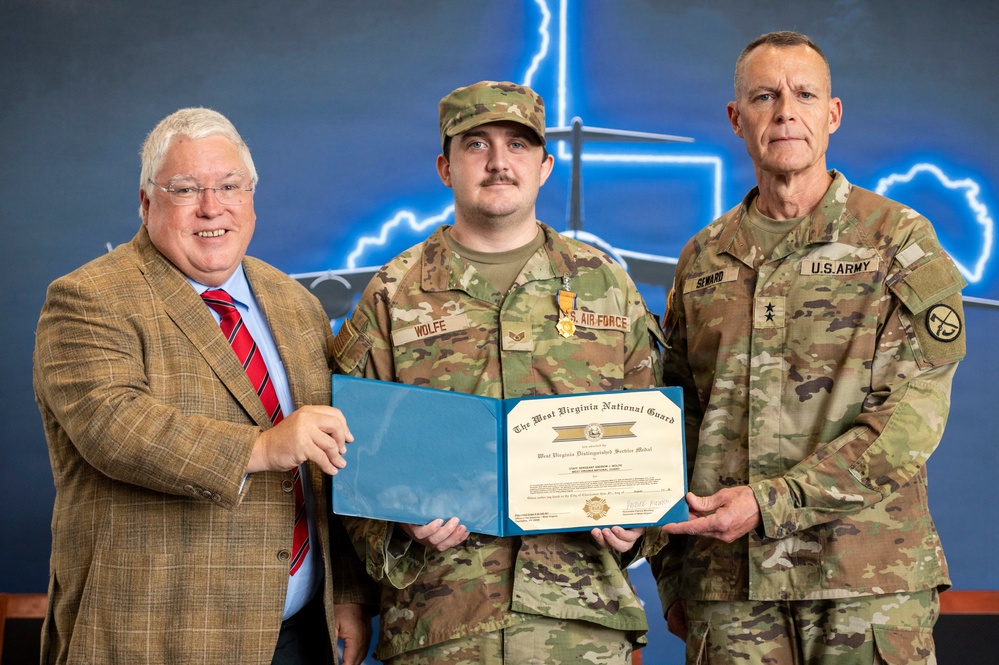 Three individuals pose for a formal military award ceremony photo. The central figure, wearing a U.S. Air Force uniform with the name tape 'WOLFE,' is holding a West Virginia National Guard certificate alongside a civilian in a tan blazer and a U.S. Army officer with the name tape 'SEWARD' bearing two-star general rank insignia. The certificate appears to be a West Virginia Distinguished Service Medal award.