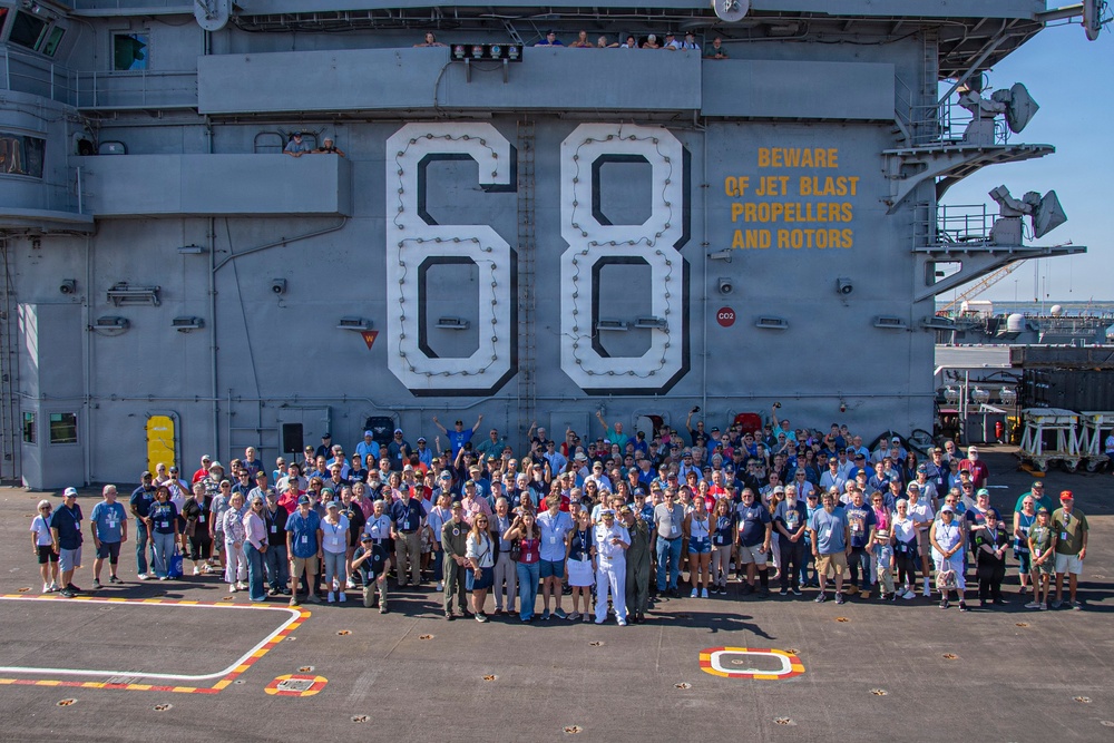 A large group of civilians and military personnel pose for a group photograph on the flight deck of USS Nimitz (CVN-68), a United States Navy aircraft carrier. The hull number '68' is prominently displayed on the island superstructure, along with a safety warning reading 'BEWARE OF JET BLAST PROPELLERS AND ROTORS' in yellow text. The event appears to be an organized tour or commemorative visit, with participants wearing lanyards/badges.