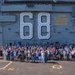 A large group of civilians and military personnel pose for a group photograph on the flight deck of USS Nimitz (CVN-68), a United States Navy aircraft carrier. The hull number '68' is prominently displayed on the island superstructure, along with a safety warning reading 'BEWARE OF JET BLAST PROPELLERS AND ROTORS' in yellow text. The event appears to be an organized tour or commemorative visit, with participants wearing lanyards/badges.
