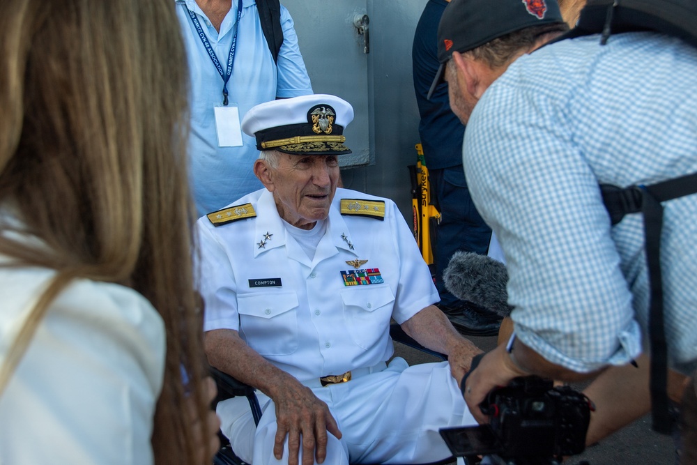 An elderly U.S. Navy officer in full dress white uniform, identified by his name tag as 'COMPTON,' is seated in a wheelchair and being interviewed by media personnel. He wears the rank insignia of a Rear Admiral (two stars on shoulder boards) and displays multiple service ribbons and an aviator badge. The scene appears to be an outdoor public or ceremonial event with press coverage.