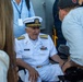 An elderly U.S. Navy officer in full dress white uniform, identified by his name tag as 'COMPTON,' is seated in a wheelchair and being interviewed by media personnel. He wears the rank insignia of a Rear Admiral (two stars on shoulder boards) and displays multiple service ribbons and an aviator badge. The scene appears to be an outdoor public or ceremonial event with press coverage.