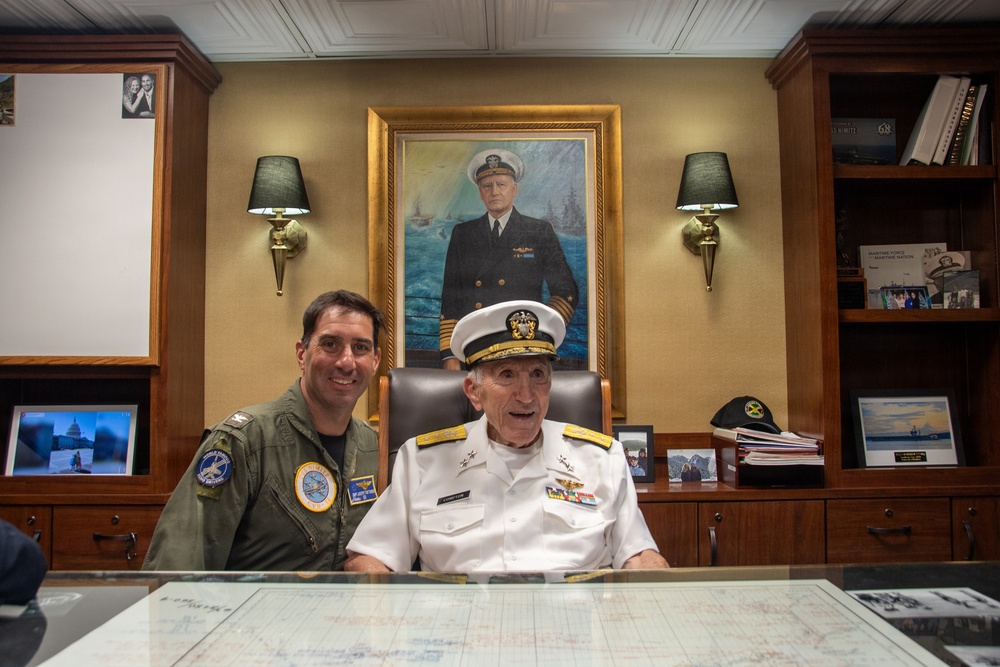 Two military personnel are posed together at a large desk in what appears to be a formal office or study. One individual is wearing a green flight suit with squadron patches, while the other is an elderly senior naval officer in white dress uniform with admiral-rank insignia. Behind them hangs a large formal oil portrait of a naval officer in dress blues.