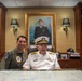 Two military personnel are posed together at a large desk in what appears to be a formal office or study. One individual is wearing a green flight suit with squadron patches, while the other is an elderly senior naval officer in white dress uniform with admiral-rank insignia. Behind them hangs a large formal oil portrait of a naval officer in dress blues.