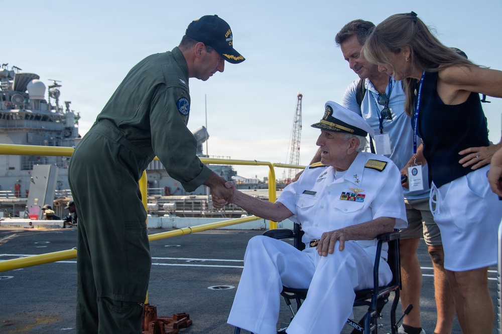 A military officer in green flight suit shakes hands with an elderly admiral in white dress uniform who is seated in a wheelchair on what appears to be a naval vessel's deck. Two civilians, a man and a woman wearing visitor badges, stand nearby observing the greeting. The setting appears to be a U.S. Navy ship at a port or naval base.