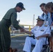 A military officer in green flight suit shakes hands with an elderly admiral in white dress uniform who is seated in a wheelchair on what appears to be a naval vessel's deck. Two civilians, a man and a woman wearing visitor badges, stand nearby observing the greeting. The setting appears to be a U.S. Navy ship at a port or naval base.