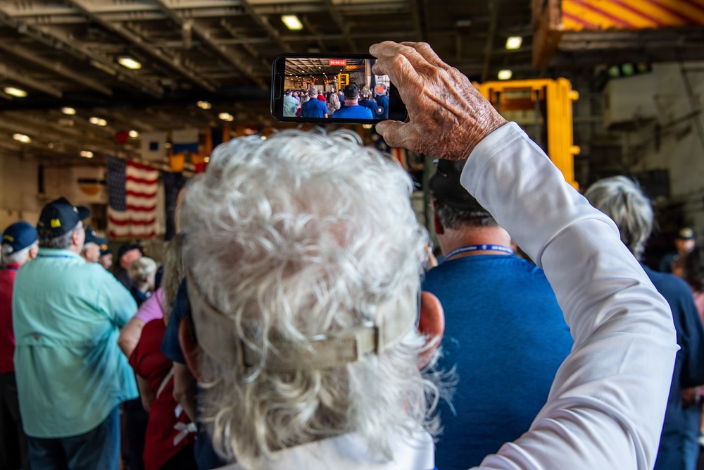 An elderly person with white curly hair holds a smartphone overhead to record or photograph a crowded indoor gathering. The event appears to be a patriotic or military ceremony, evidenced by an American flag and attendees wearing military-style caps in the background. The setting appears to be inside a large industrial or naval vessel hangar bay.