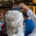 An elderly person with white curly hair holds a smartphone overhead to record or photograph a crowded indoor gathering. The event appears to be a patriotic or military ceremony, evidenced by an American flag and attendees wearing military-style caps in the background. The setting appears to be inside a large industrial or naval vessel hangar bay.