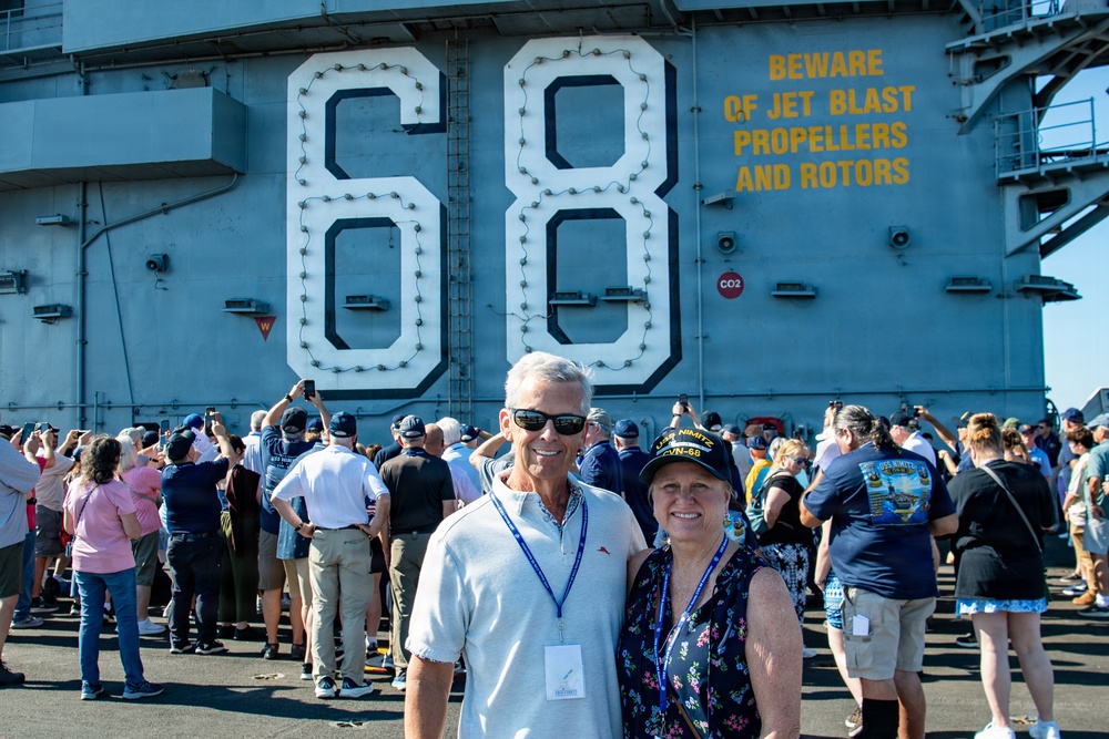 Two individuals pose for a photo on the flight deck of USS Nimitz (CVN-68), a United States Navy aircraft carrier. The woman is wearing an official USS Nimitz CVN-68 cap, and both wear blue lanyards with visitor badges. A crowd of visitors surrounds them, many taking photographs of the large hull number '68' and the safety warning painted on the ship's island structure.