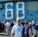 Two individuals pose for a photo on the flight deck of USS Nimitz (CVN-68), a United States Navy aircraft carrier. The woman is wearing an official USS Nimitz CVN-68 cap, and both wear blue lanyards with visitor badges. A crowd of visitors surrounds them, many taking photographs of the large hull number '68' and the safety warning painted on the ship's island structure.