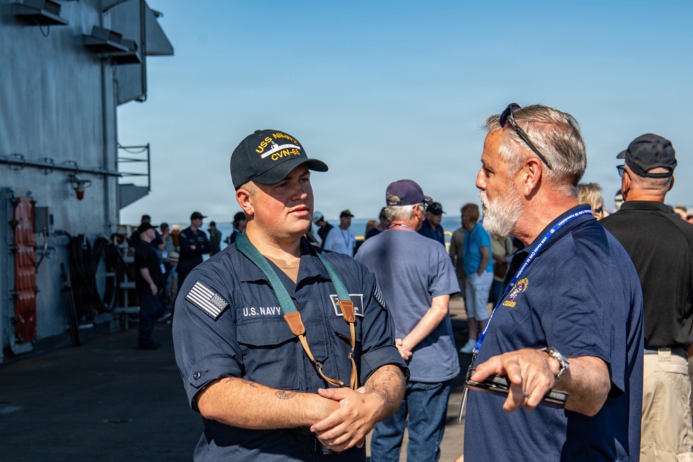A U.S. Navy sailor in uniform is engaged in conversation with a civilian man wearing a blue polo shirt and a lanyard on what appears to be the deck of a naval vessel. The sailor is wearing a cap marked 'USS' and 'CVN-6X' and a U.S. Navy uniform with green suspenders. A group of civilians and personnel are visible in the background, suggesting a public tour or community outreach event aboard the ship.