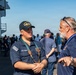 A U.S. Navy sailor in uniform is engaged in conversation with a civilian man wearing a blue polo shirt and a lanyard on what appears to be the deck of a naval vessel. The sailor is wearing a cap marked 'USS' and 'CVN-6X' and a U.S. Navy uniform with green suspenders. A group of civilians and personnel are visible in the background, suggesting a public tour or community outreach event aboard the ship.